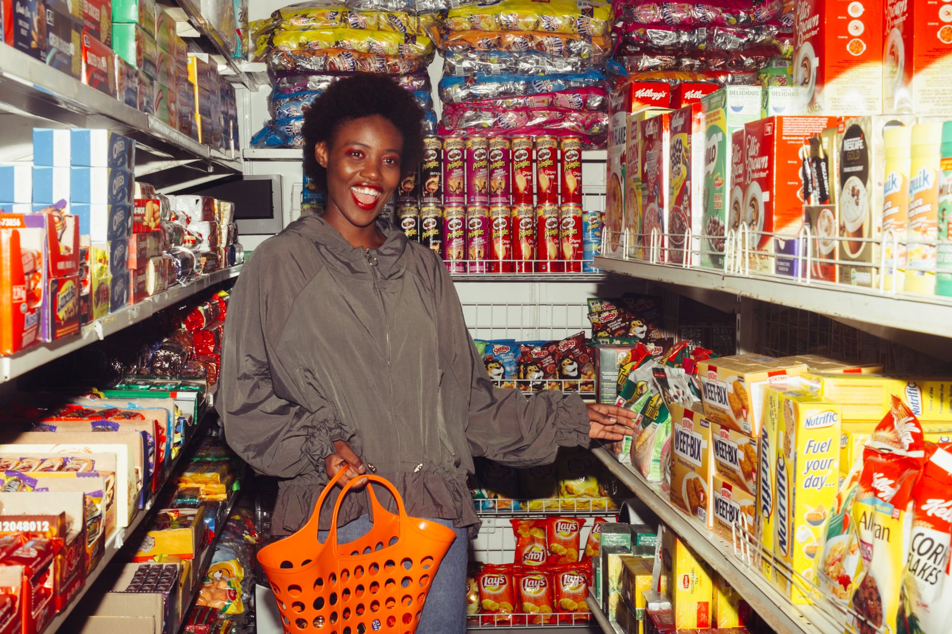 Happy African American lady in casual outfit with shopping basket in hand choosing food from products in small supermarket among colorful boxes on shelves and smiling with mouth opened
